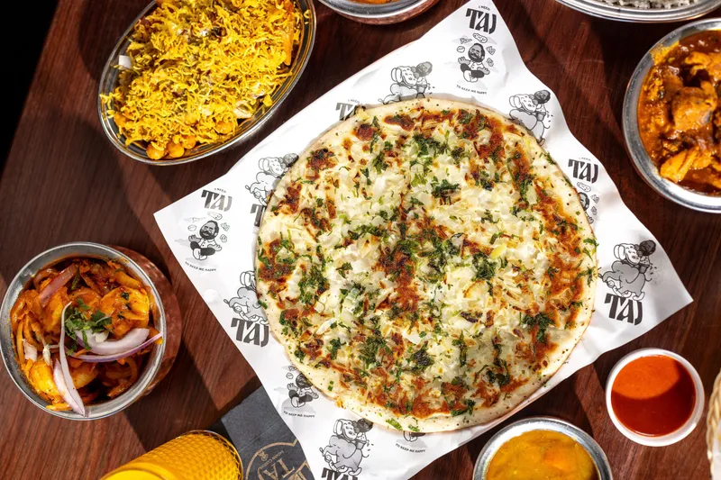 Overhead spread at Taj Indian Cuisine in South Portland — an onion-and-herb uttapam on Taj-branded paper, surrounded by biryani, chicken curry, a shrimp dish, dal and chutneys on a wood table
