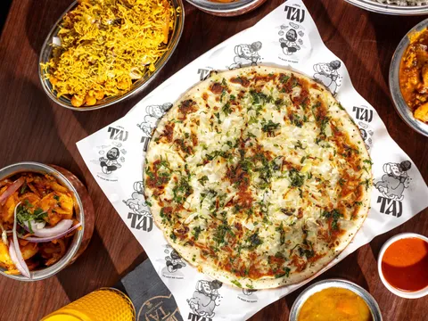 Overhead spread at Taj Indian Cuisine in South Portland — an onion-and-herb uttapam on Taj-branded paper, surrounded by biryani, chicken curry, a shrimp dish, dal and chutneys on a wood table