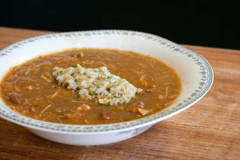 A bowl of gumbo topped with a scoop of white rice and chopped chives, served in a patterned-rim white bowl on a wooden table at Virtue in Chicago