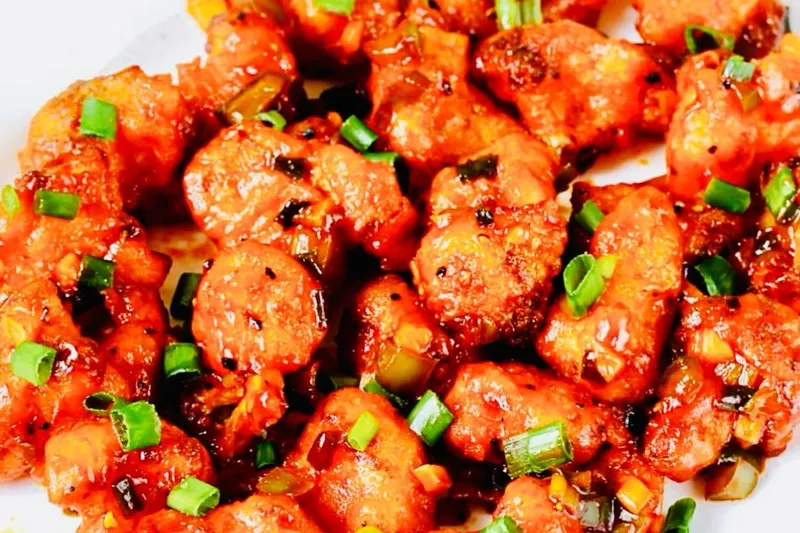 A plate of Rasam's Indo-Chinese style battered chicken tossed in a glossy red chili-garlic sauce and garnished with scallions, on a floral-rimmed plate