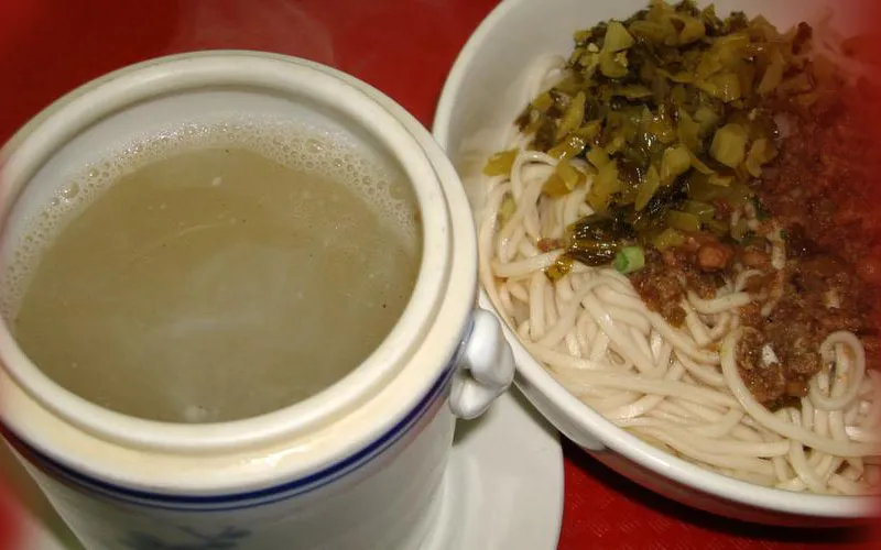 Taiwan Pork Chop House in New York — a lidded ceramic pot of clear herbal soup beside a bowl of dry lo mein topped with pickled mustard greens and minced pork