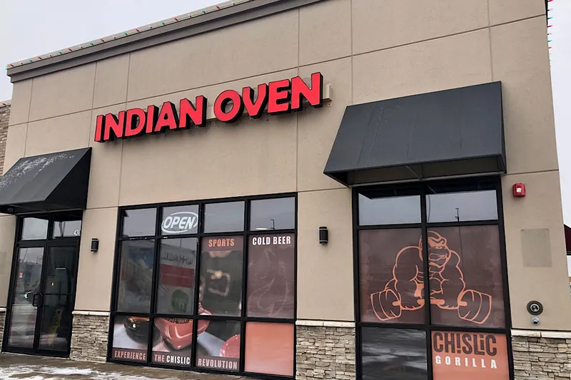 Exterior of Indian Oven in Sioux Falls, a tan strip-mall storefront with red INDIAN OVEN channel-letter signage, black awnings and an OPEN sign in the window