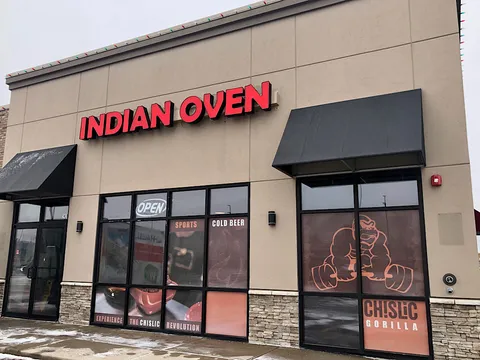 Exterior of Indian Oven in Sioux Falls, a tan strip-mall storefront with red INDIAN OVEN channel-letter signage, black awnings and an OPEN sign in the window