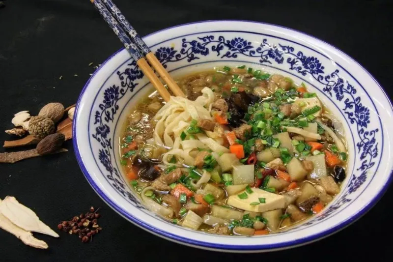 Original Pork Noodles at Shan Xi Magic Kitchen — hand-made noodles in broth with diced tofu, carrot, wood ear and scallion in a blue-and-white porcelain bowl, dried Chinese spices arranged alongside