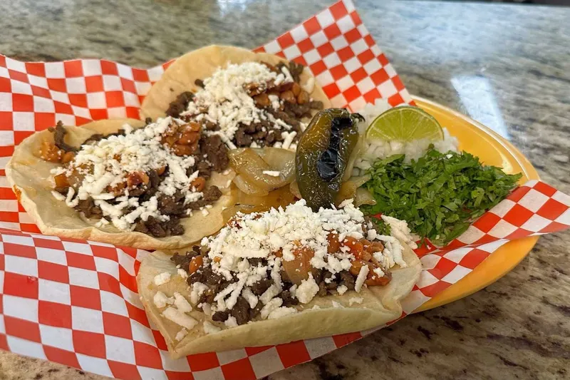 Three open-faced street tacos topped with chopped steak and cotija cheese, with a grilled jalapeño, lime, cilantro and onion in a checkered basket at Taqueria Datapoint