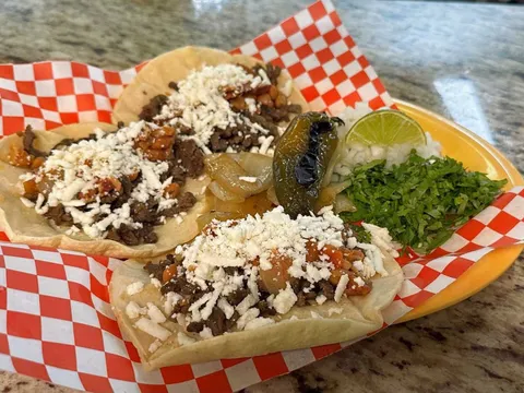 Three open-faced street tacos topped with chopped steak and cotija cheese, with a grilled jalapeño, lime, cilantro and onion in a checkered basket at Taqueria Datapoint