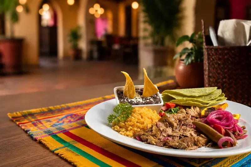 Plated Oaxacan dish of shredded meat with yellow rice, black beans, pickled onions and tortillas on a colorful woven table runner, with the warmly lit arched dining room of Fiesta Oaxaca behind
