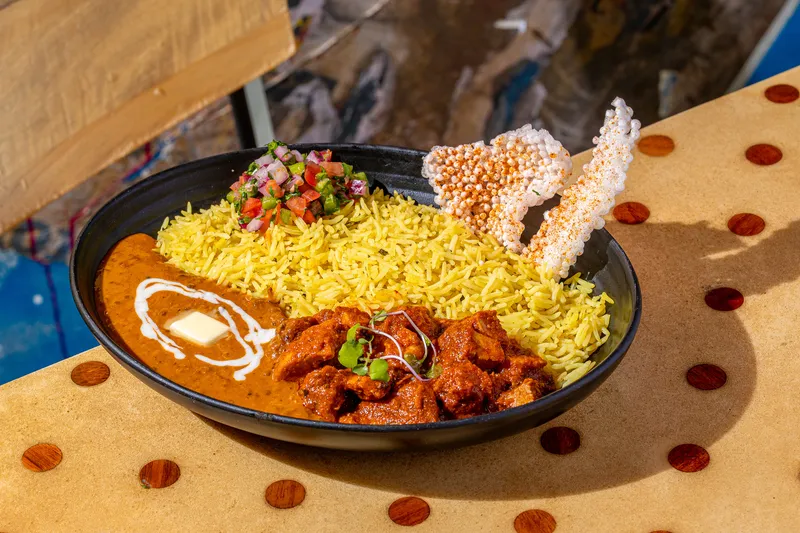 A Curry Up Now bowl of butter chicken makhni with turmeric rice, pico kachumber and puffed sabudana papad in a black bowl, brand food photograph