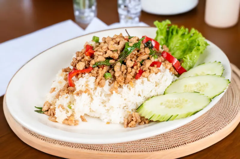 Thai basil chicken with red chilis over jasmine rice with cucumber and lettuce on a white plate at Lers Ros Thai