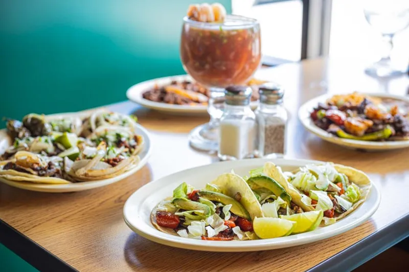 Plates of tacos with avocado and lettuce alongside a shrimp cocktail on a table at Taqueria Los Compadres