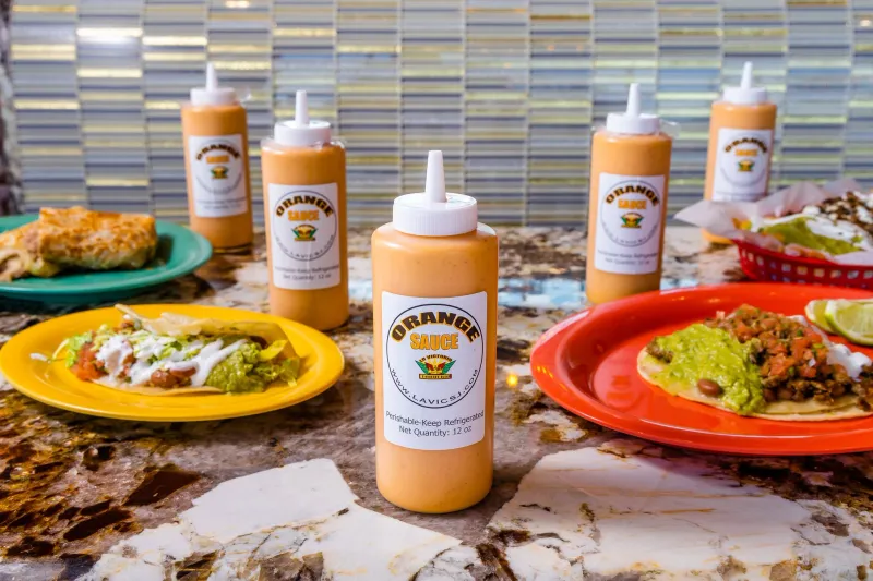 Bottles of La Victoria Taqueria's signature Orange Sauce lined up on a marble counter beside plates of tacos
