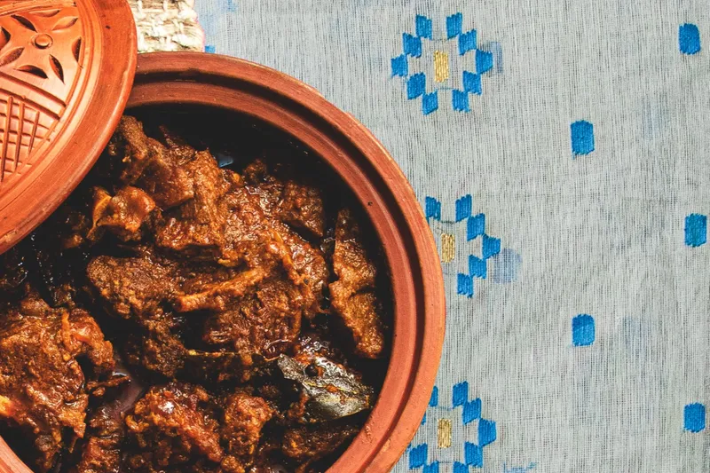Kosha mangsho-style goat curry served in a lidded terracotta clay pot on a blue-and-gold jamdani-patterned cloth at Korai Kitchen in Jersey City