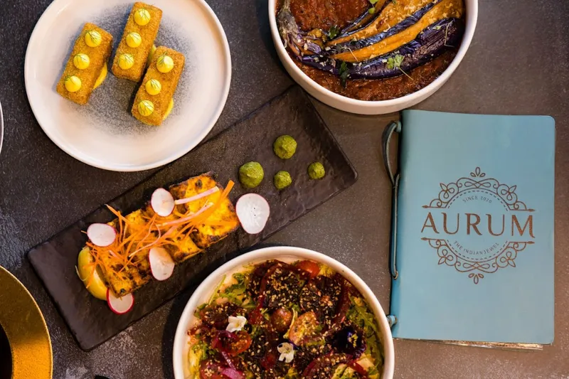 A spread of Aurum's modern Indian dishes — paneer tikka, kofta, eggplant curry and a chaat bowl — beside the branded Aurum menu booklet