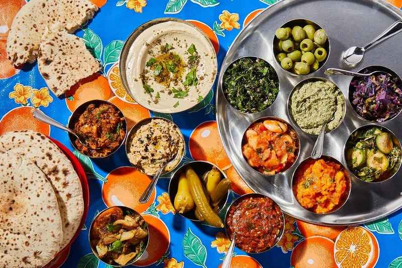 Overhead spread of Laser Wolf salatim - hummus, olives, dips and pita on a colorful tablecloth