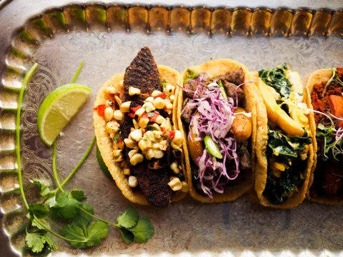 A row of Zona Rosa tacos — blackened bass, hanger steak, and more — arranged on an engraved silver tray with lime and cilantro