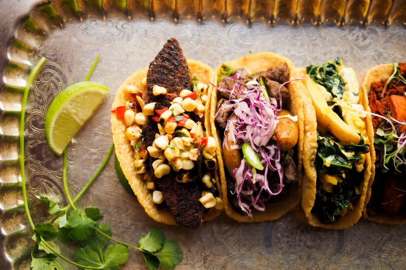 A row of Zona Rosa tacos — blackened bass, hanger steak, and more — arranged on an engraved silver tray with lime and cilantro