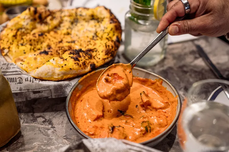 A spoon lifting creamy butter-chicken-style curry from a steel bowl, with charred garlic naan alongside, from Bombay Blue Indian Bistro in Edison, New Jersey
