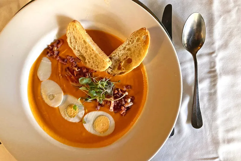 A chilled tomato soup (salmorejo) garnished with microgreens, chopped chorizo and toasted bread on a white-clothed table at Madeira Restaurant