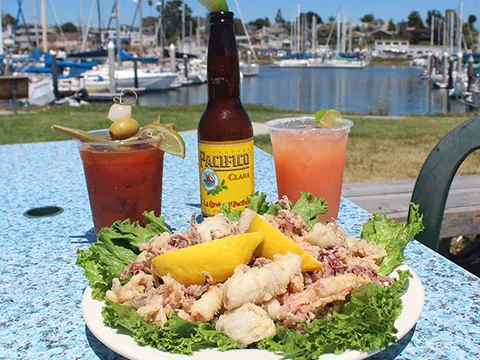 A plate of fried calamari with lemon and lettuce beside a beer and two cocktails on a patio table at Aldo's Harbor Restaurant, with the Santa Cruz Harbor and moored sailboats behind