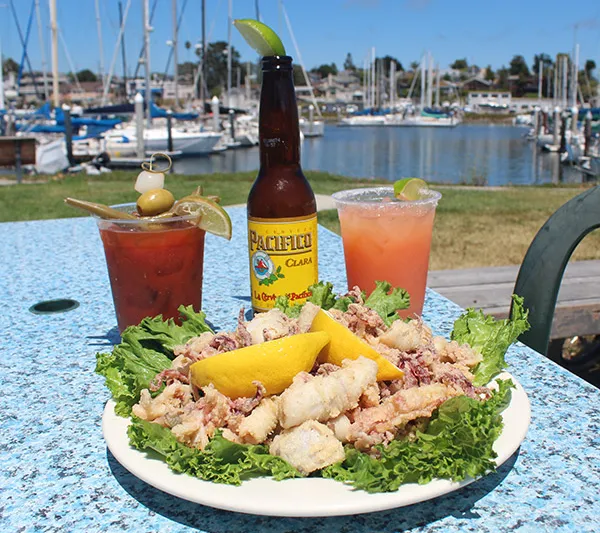 A plate of fried calamari with lemon and lettuce beside a beer and two cocktails on a patio table at Aldo's Harbor Restaurant, with the Santa Cruz Harbor and moored sailboats behind