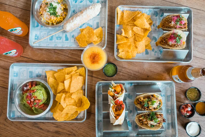 Overhead spread of tacos, tortilla chips, guacamole, elote and salsas on metal trays at Coyo Taco