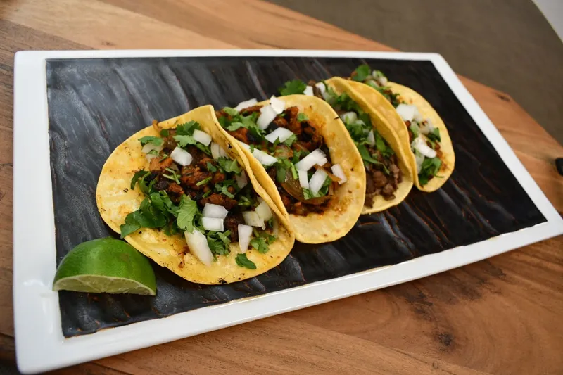 A plate of four Basilios Tacos street tacos — soft corn tortillas topped with grilled meat, chopped onion and cilantro — with a lime wedge, on a dark rectangular plate over a wood table