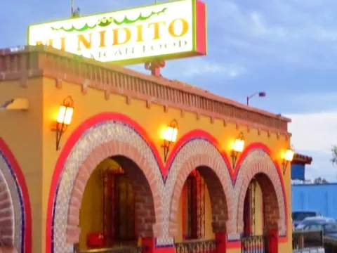 The illuminated 'Mi Nidito Mexican Food' rooftop sign above the colorful yellow, red and tiled arched storefront of Mi Nidito Restaurant in South Tucson at dusk