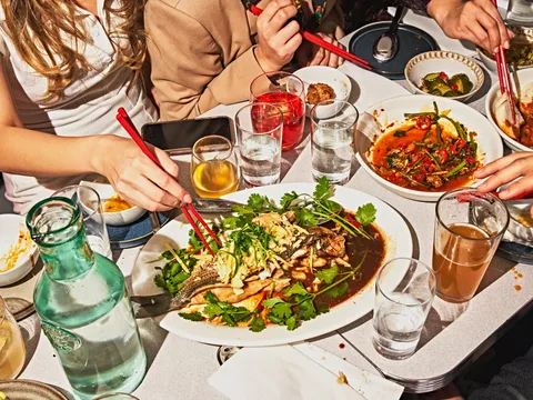 A shared table at Wenwen in Brooklyn — a whole steamed striped bass in black-sugar yuzu sauce with cilantro, surrounded by stir-fried dishes, pickles and glasses of tea (diners' hands with chopsticks in frame, no faces)