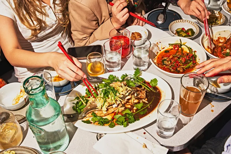A shared table at Wenwen in Brooklyn — a whole steamed striped bass in black-sugar yuzu sauce with cilantro, surrounded by stir-fried dishes, pickles and glasses of tea (diners' hands with chopsticks in frame, no faces)