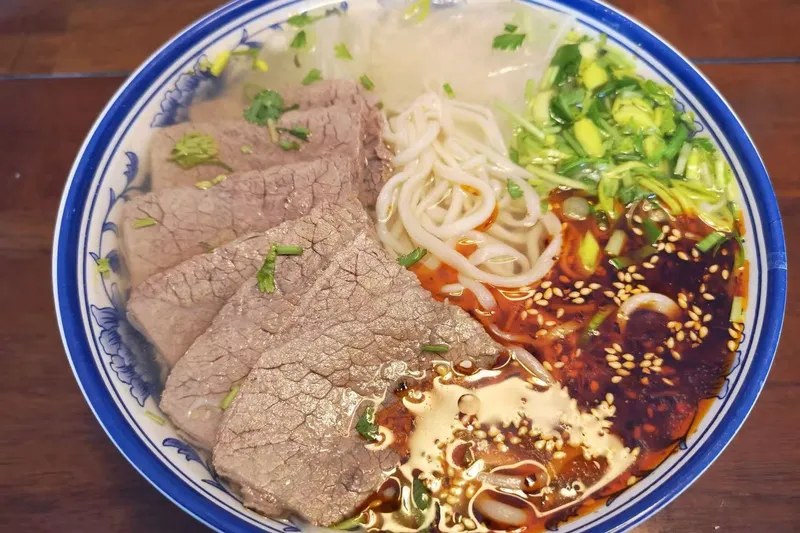 A bowl of Kung Fu Noodle's Classic Lanzhou beef noodle soup with sliced beef, hand-pulled noodles, cilantro, garlic greens and chili oil in a blue-and-white porcelain bowl