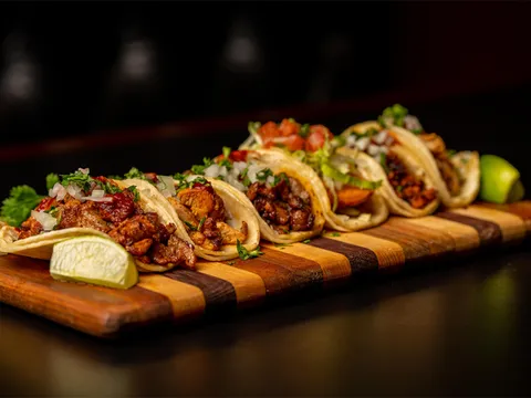 A row of five street tacos on soft corn tortillas — topped with grilled meats, onion, cilantro and salsa — plated on a striped wooden board with lime wedges at Sin Fronteras Tacos