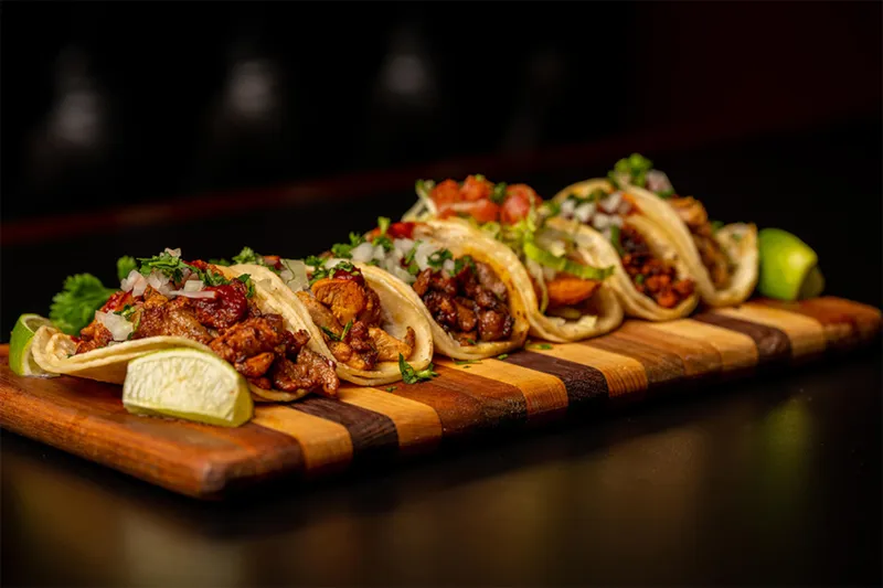 A row of five street tacos on soft corn tortillas — topped with grilled meats, onion, cilantro and salsa — plated on a striped wooden board with lime wedges at Sin Fronteras Tacos