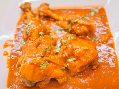 Bone-in chicken curry in a rich orange-red gravy garnished with cilantro on a white plate at Planet Bombay