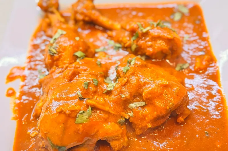 Bone-in chicken curry in a rich orange-red gravy garnished with cilantro on a white plate at Planet Bombay