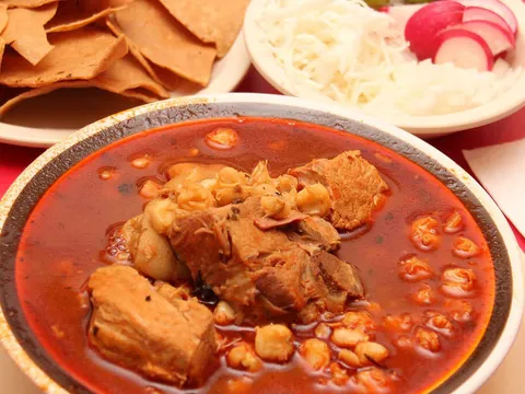 A bowl of pozole with hominy and pork in red broth at Sabores Oaxaqueños Restaurant, served with a plate of fried tortilla chips and a side of shredded cabbage, radishes and onion