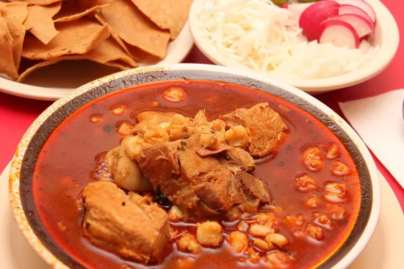 A bowl of pozole with hominy and pork in red broth at Sabores Oaxaqueños Restaurant, served with a plate of fried tortilla chips and a side of shredded cabbage, radishes and onion