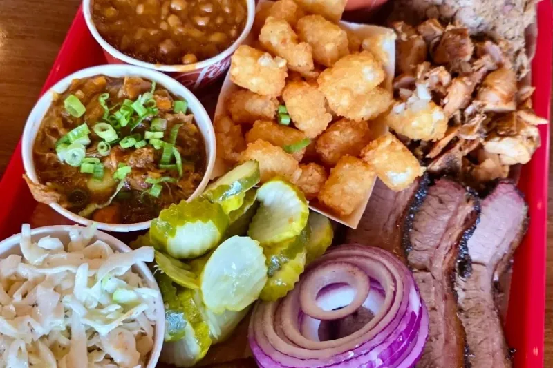 Barbecue platter on a red tray at Feast BBQ — sliced brisket, pulled pork, smoked sausage, tater tots, baked beans, burgoo, coleslaw, pickles and red onion