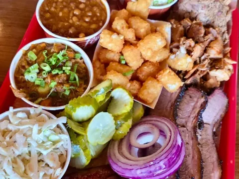 Barbecue platter on a red tray at Feast BBQ — sliced brisket, pulled pork, smoked sausage, tater tots, baked beans, burgoo, coleslaw, pickles and red onion