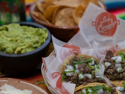 Basket of carne asada street tacos on Los Comales branded paper alongside guacamole, refried beans with melted cheese, Mexican rice and tortilla chips