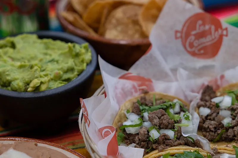 Basket of carne asada street tacos on Los Comales branded paper alongside guacamole, refried beans with melted cheese, Mexican rice and tortilla chips