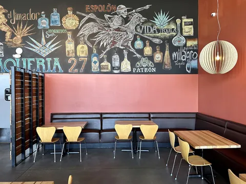 The dining room at Taqueria 27 in Salt Lake City, with a chalkboard mural reading "Taqueria 27" surrounded by illustrated tequila and mezcal bottles above a leather banquette and pale wood chairs