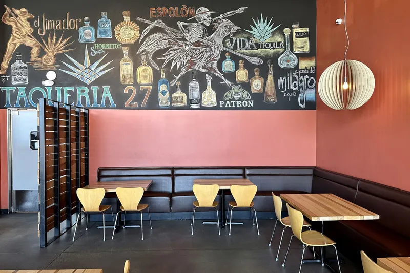The dining room at Taqueria 27 in Salt Lake City, with a chalkboard mural reading "Taqueria 27" surrounded by illustrated tequila and mezcal bottles above a leather banquette and pale wood chairs