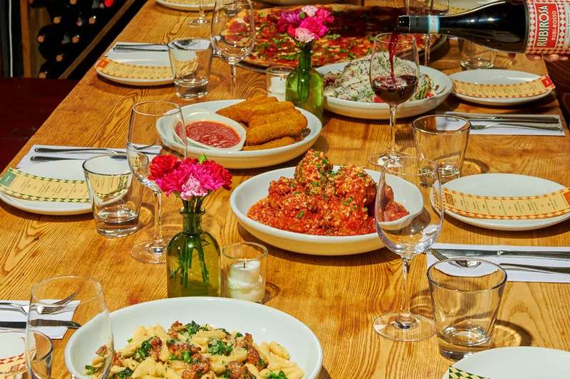 Family-style spread at Rubirosa with the signature tie-dye pizza, cavatelli, meatballs and a bottle of Rubi Rosso wine being poured on a wooden table