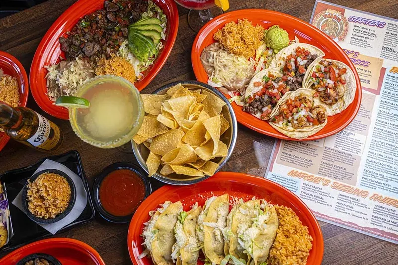 Overhead spread of Rudy's Tenampa Taqueria dishes on red plates — street tacos with pico de gallo, a carne asada platter, chips, salsa and margaritas, beside the restaurant's printed menu