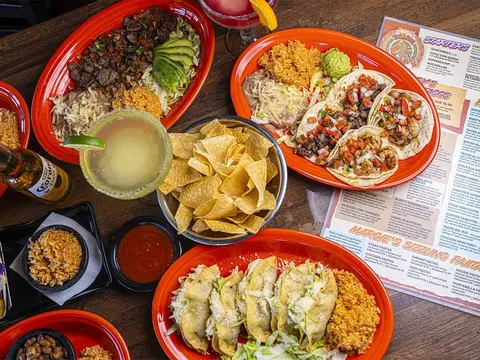 Overhead spread of Rudy's Tenampa Taqueria dishes on red plates — street tacos with pico de gallo, a carne asada platter, chips, salsa and margaritas, beside the restaurant's printed menu