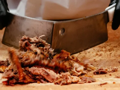 Gloved hands chopping wood-cooked whole-hog barbecue with a cleaver on a board at Skylight Inn BBQ in Ayden, North Carolina