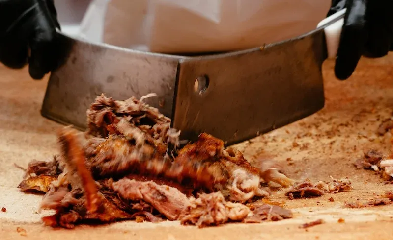 Gloved hands chopping wood-cooked whole-hog barbecue with a cleaver on a board at Skylight Inn BBQ in Ayden, North Carolina