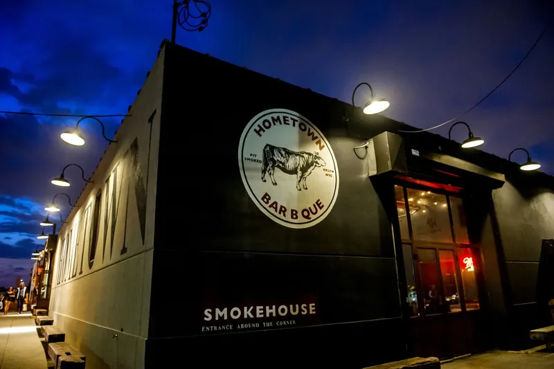 Exterior of Hometown Bar-B-Que at dusk, a black building at 454 Van Brunt Street in Red Hook, Brooklyn, with the round Hometown Bar B Que steer logo and string lights