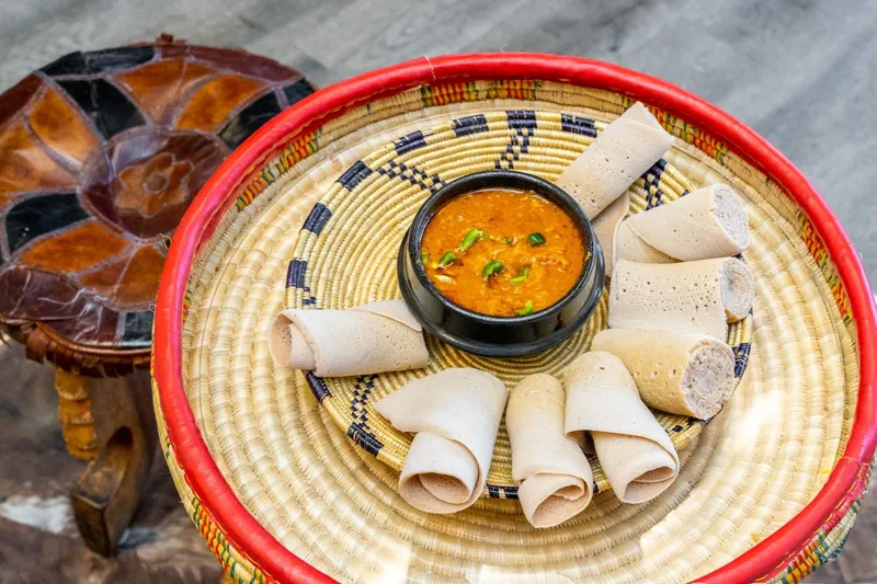 Rolls of injera and a spiced stew served in a woven mesob basket beside a traditional leather stool at Zeni Ethiopian Restaurant, San Jose