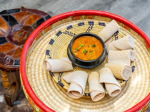 Rolls of injera and a spiced stew served in a woven mesob basket beside a traditional leather stool at Zeni Ethiopian Restaurant, San Jose